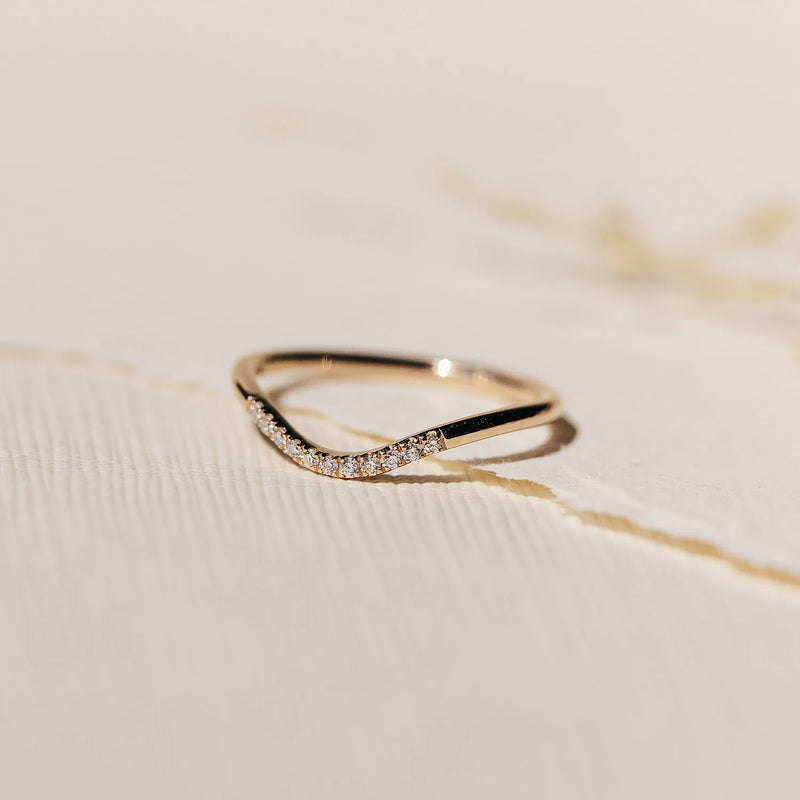 A delicate wave eternity ring by beloved by bluboho lies on a textured, off-white surface. Crafted in 14k yellow gold, the ring features a slim band that gently curves, adorned with a small cluster of sparkling white diamonds on one side, adding a touch of elegance and sophistication to its minimalist design.